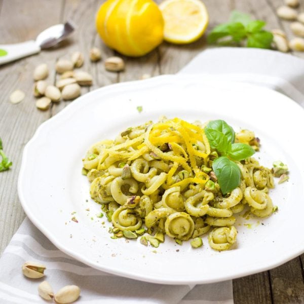 a dish of pasta with pesto, a sprig of basil placed on a white dish surrounded by lemon and pistachios.